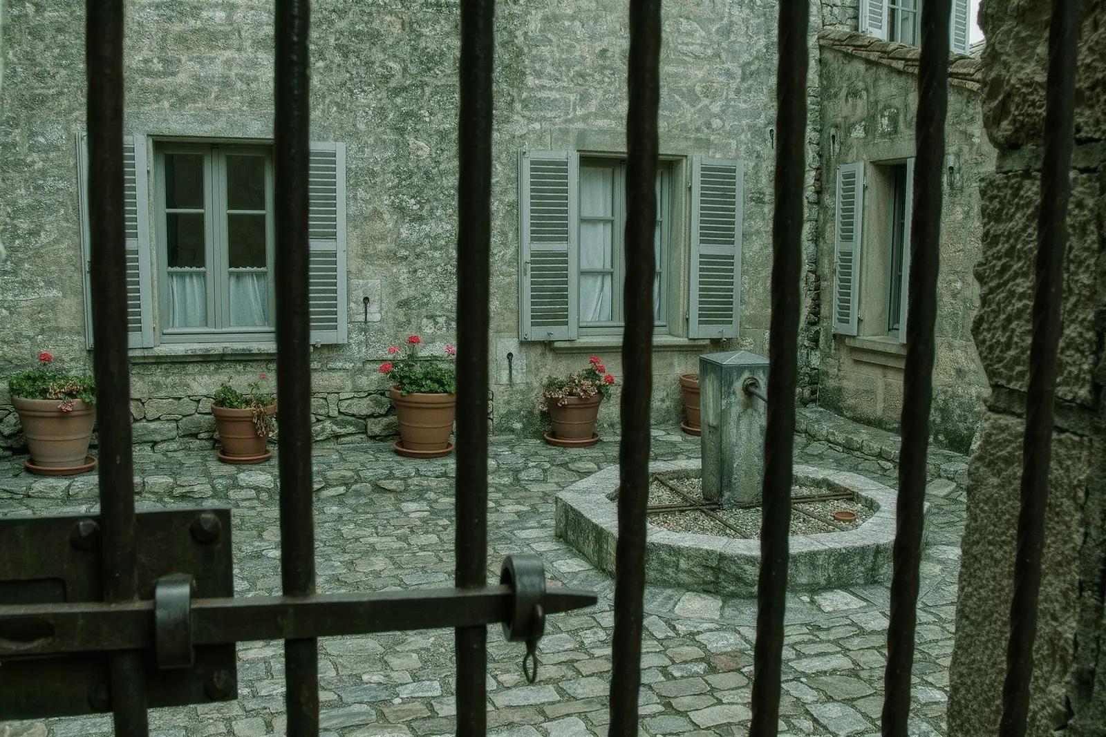 The stone courtyard of an inland Mallorcan finca with shuttered windows, a dry fountain and terracotta pots of tired geraniums beside a worn iron gate.
