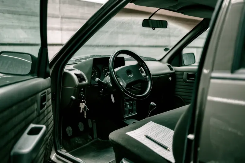A view across the open driver's door of a small hire car in an airport car park, the dashboard and fuel gauge visible, a paper condition report and pen on the seat.