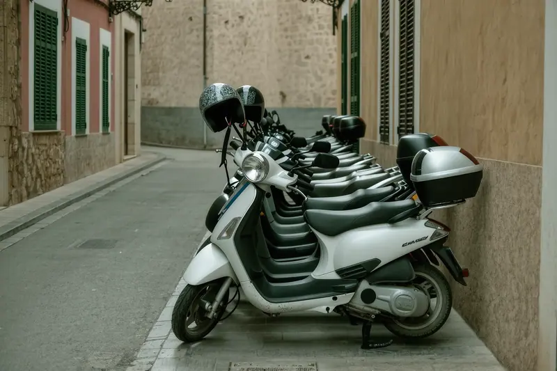 A row of small scooters in a rental rack on a narrow street in Alcúdia old town, helmets hanging on the mirrors and a shuttered shopfront behind.