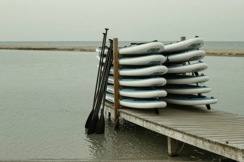 Paddleboards stacked on a wooden rack beside a pontoon in the shallow water of a wide sheltered bay, flat sand and low pine behind.