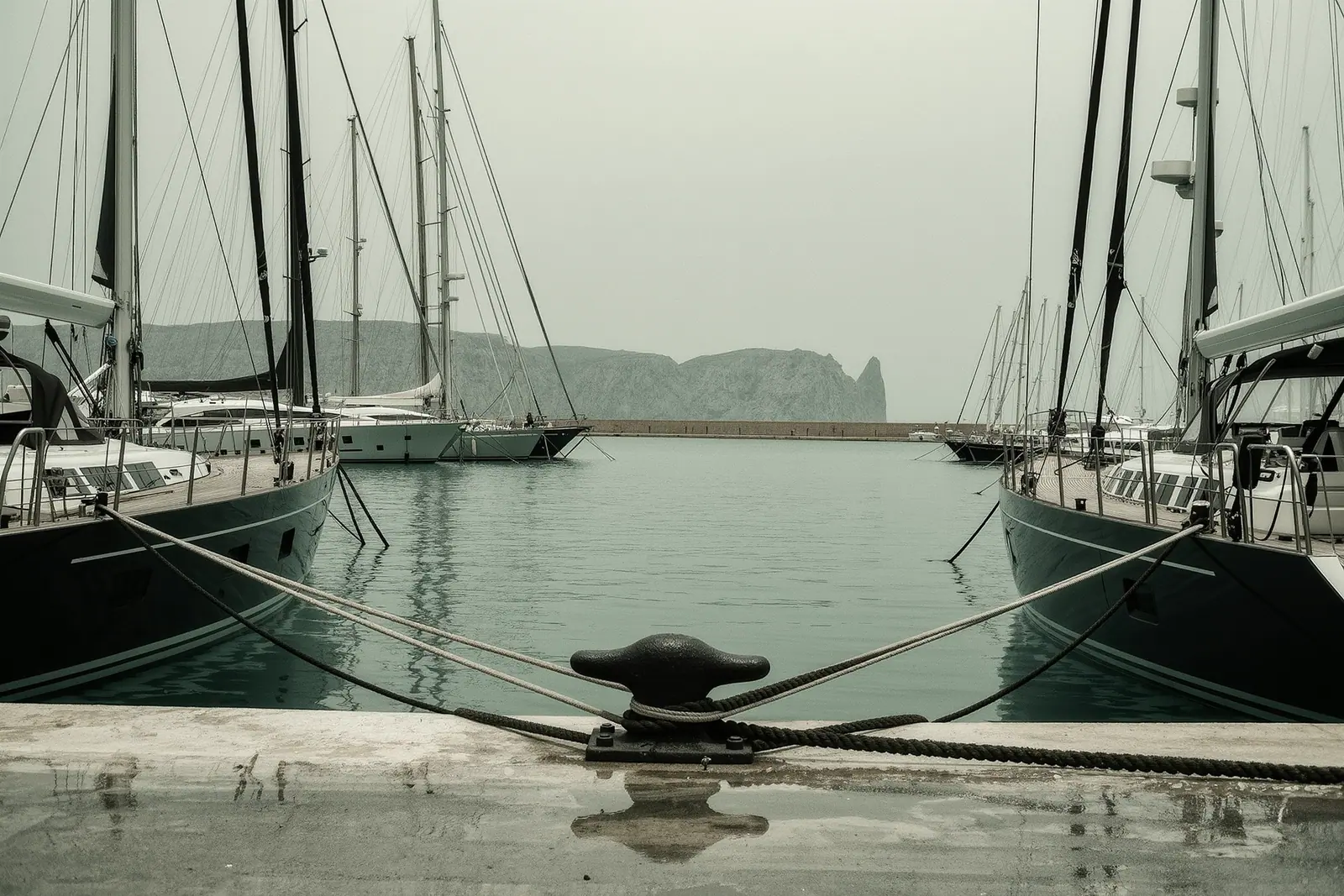 Charter sailing yachts moored stern-to along a working quay in Palma, mooring lines and a cast-iron cleat in the foreground, the grey Tramuntana ridge behind the masts.