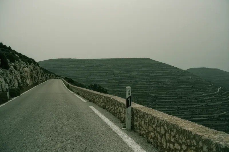 An empty stretch of the Ma-10 mountain road with a dry stone wall alongside and a cycling route marker post, no traffic in sight.
