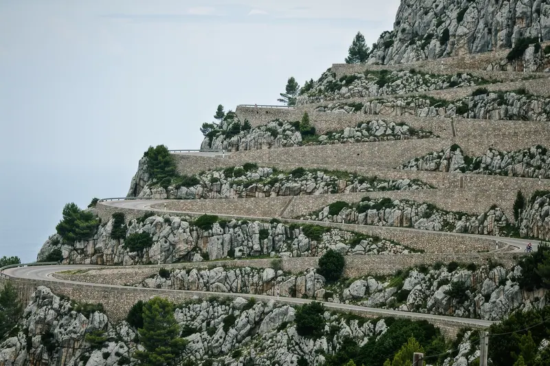 Stacked hairpin bends of a narrow limestone road descending a steep slope in the Serra de Tramuntana, dry stone walls on the outside of the bends.